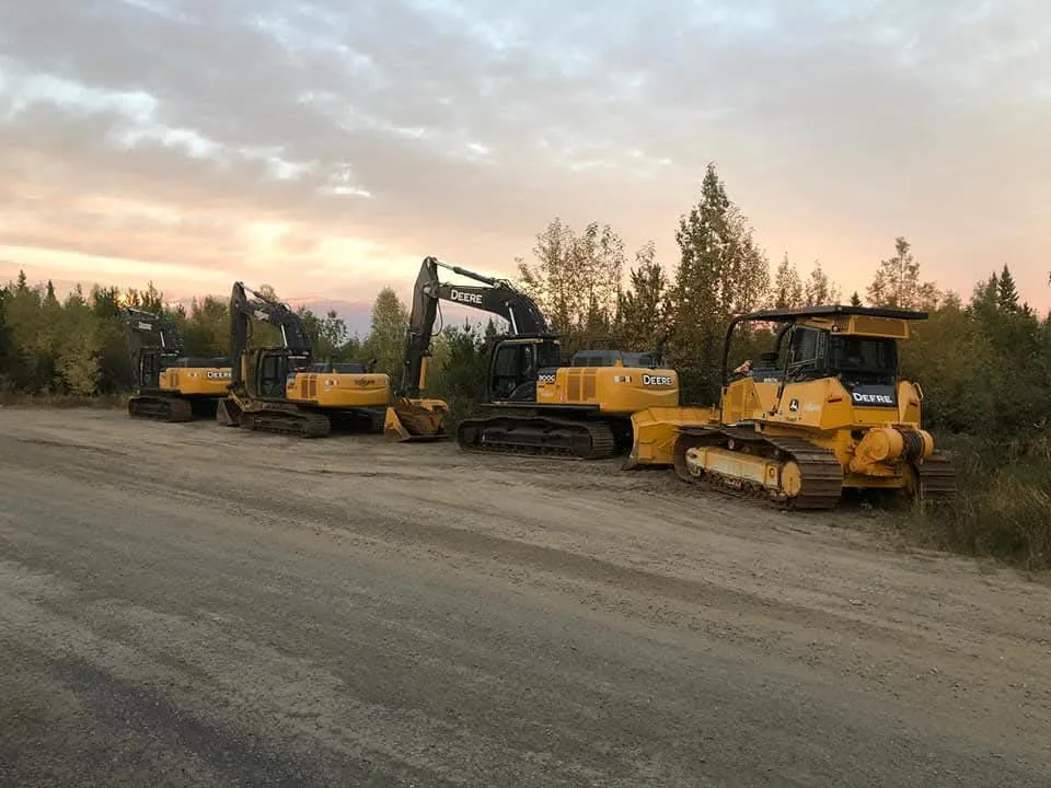 Excavators and a dozer lined up at the edge of a clearing at dusk