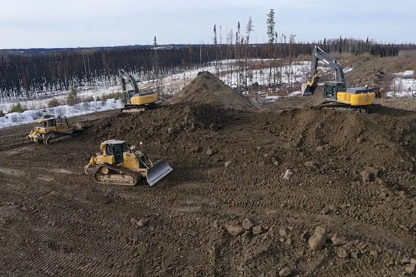 Two excavators and two dozers moving soil beside burnt standing timber