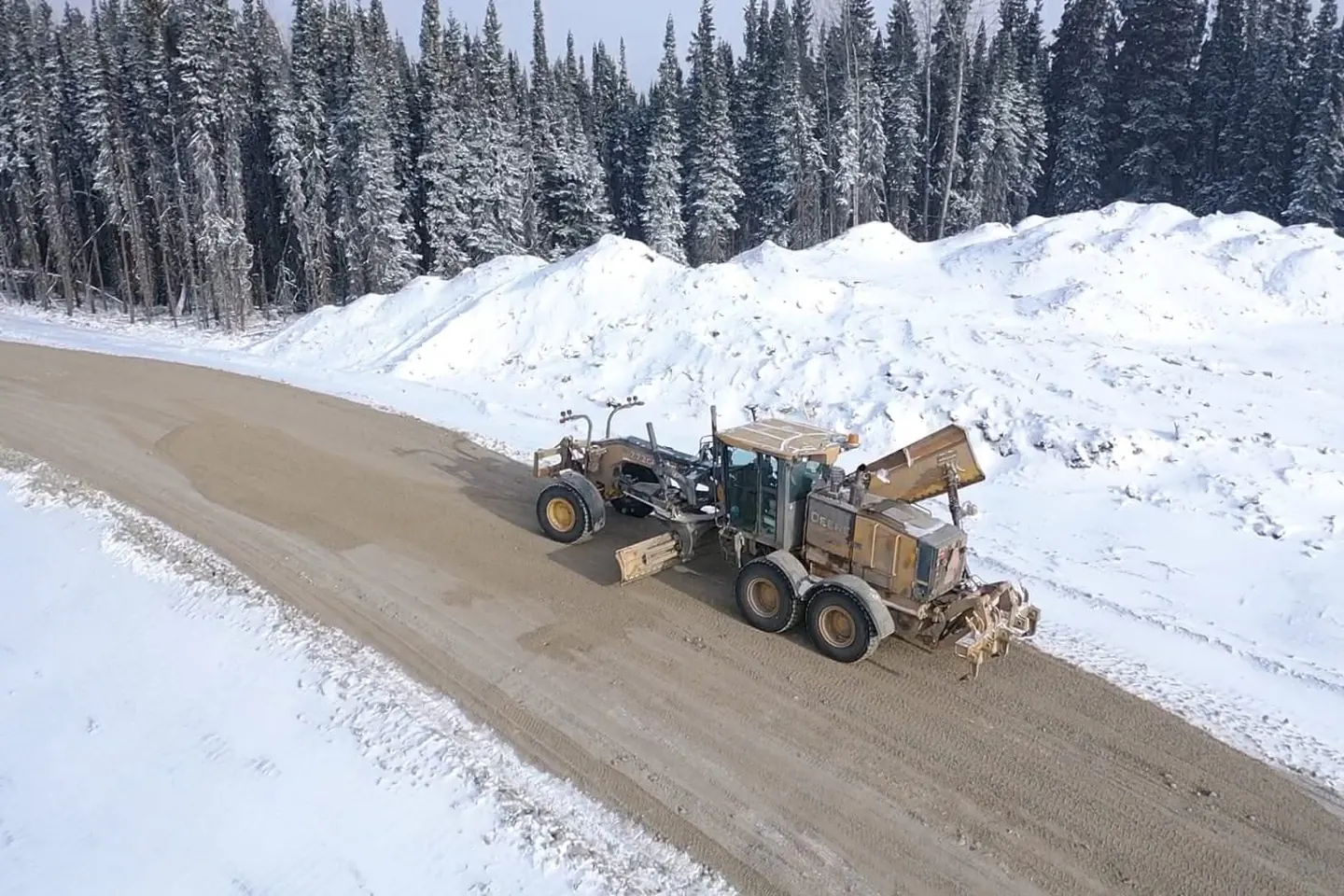 Motor grader shaping a gravel road bordered by snow banks